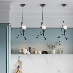 K&ouml;nnte beinhalten: Drei schwarze Pendelleuchten mit einem einzigartigen Design. Jede Leuchte zeigt eine stilisierte Figur, die eine runde, wei&szlig;e Gl&uuml;hbirne h&auml;lt. Die Figuren scheinen sich zu bewegen, mit ausgestreckten Armen und Beinen. Die Leuchten h&auml;ngen von der Decke in einer modernen K&uuml;che.