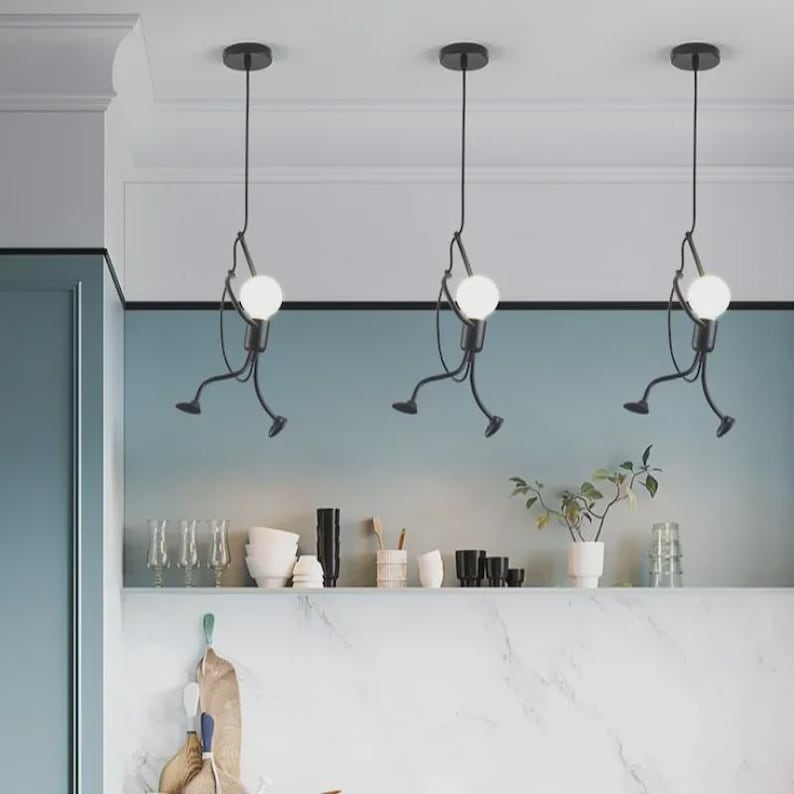 K&ouml;nnte beinhalten: Drei schwarze Pendelleuchten mit einem einzigartigen Design. Jede Leuchte zeigt eine stilisierte Figur, die eine runde, wei&szlig;e Gl&uuml;hbirne h&auml;lt. Die Figuren scheinen sich zu bewegen, mit ausgestreckten Armen und Beinen. Die Leuchten h&auml;ngen von der Decke in einer modernen K&uuml;che.