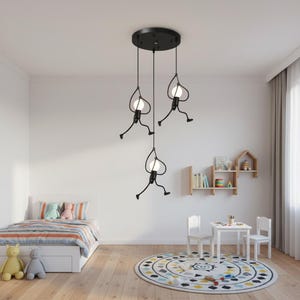 K&ouml;nnte beinhalten: Eine schwarze Pendelleuchte mit drei Lichtern und einem skurrilen Design. Jedes Licht hat eine stilisierte Figur mit einer runden Gl&uuml;hbirne, die von einer kreisf&ouml;rmigen Deckenhalterung abgeh&auml;ngt ist. Die Lampe befindet sich in einem Kinderzimmer.