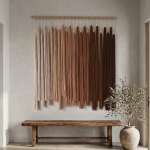 Puede incluir: Un tapiz de pared con largos hilos verticales suspendidos de una varilla de madera horizontal. El hilo muestra una paleta de colores en degradado que transita desde el crema claro y beige a la izquierda hasta el canela, &oacute;xido y marr&oacute;n oscuro.