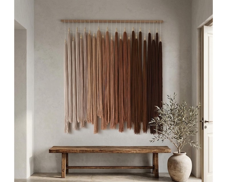 Puede incluir: Un tapiz de pared con largos hilos verticales suspendidos de una varilla de madera horizontal. El hilo muestra una paleta de colores en degradado que transita desde el crema claro y beige a la izquierda hasta el canela, &oacute;xido y marr&oacute;n oscuro.