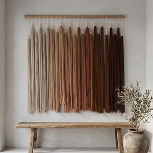 Puede incluir: Un tapiz de pared que presenta una varilla de madera horizontal con numerosas borlas de fibra largas y verticales suspendidas. Las borlas est&aacute;n dispuestas en un degradado de color, desde beige claro y tostado hasta tonos marrones c&aacute;lidos y chocolate.