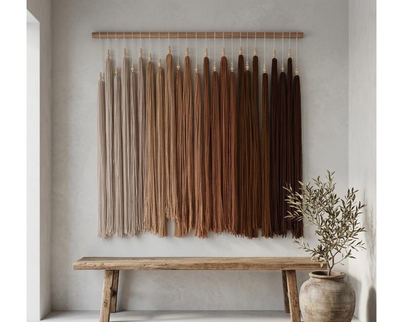 Puede incluir: Un tapiz de pared que presenta una varilla de madera horizontal con numerosas borlas de fibra largas y verticales suspendidas. Las borlas est&aacute;n dispuestas en un degradado de color, desde beige claro y tostado hasta tonos marrones c&aacute;lidos y chocolate.