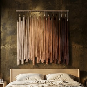 Puede incluir: Un tapiz de pared que presenta una varilla de madera horizontal con numerosas borlas de hilo largas y verticales suspendidas. Las borlas presentan un degradado de color desde crema y beige claro a la izquierda hasta marr&oacute;n oscuro a la derecha.