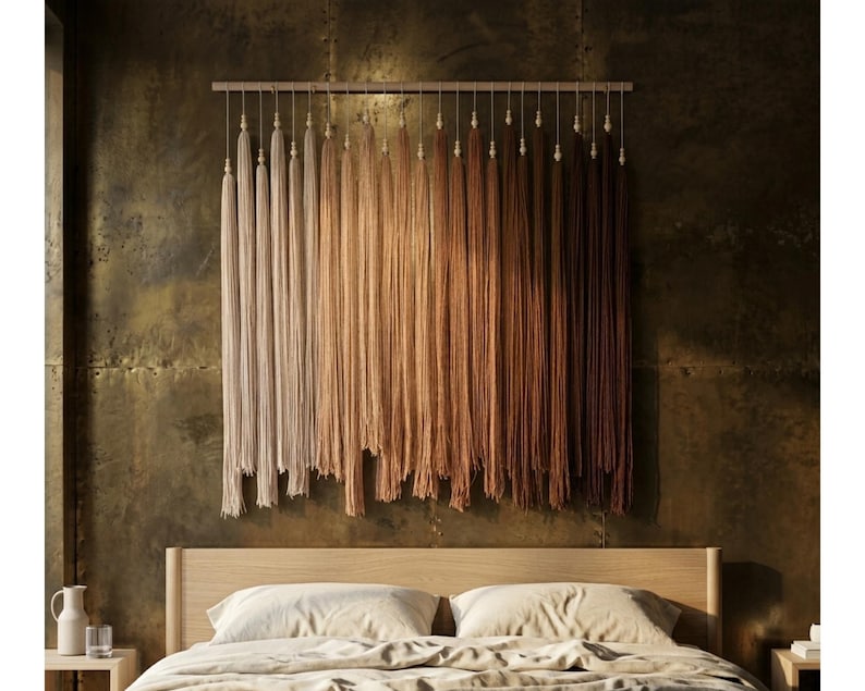 Puede incluir: Un tapiz de pared que presenta una varilla de madera horizontal con numerosas borlas de hilo largas y verticales suspendidas. Las borlas presentan un degradado de color desde crema y beige claro a la izquierda hasta marr&oacute;n oscuro a la derecha.