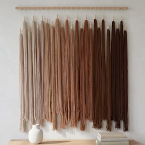 Puede incluir: Un tapiz de pared que presenta una varilla de madera horizontal con m&uacute;ltiples borlas de hilo verticales suspendidas. Las borlas est&aacute;n dispuestas en un degradado de color, pasando de beige claro y tostado a varios tonos de marr&oacute;n y chocolate oscuro.