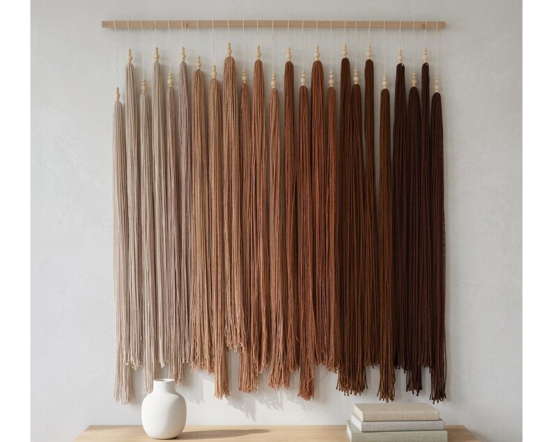 Puede incluir: Un tapiz de pared que presenta una varilla de madera horizontal con m&uacute;ltiples borlas de hilo verticales suspendidas. Las borlas est&aacute;n dispuestas en un degradado de color, pasando de beige claro y tostado a varios tonos de marr&oacute;n y chocolate oscuro.