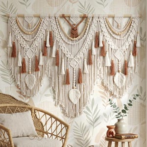 May include: A large macrame wall hanging with a wooden rod, featuring intricate woven patterns, tassels, and leaf-shaped details. The color palette includes cream, brown, and beige, creating a bohemian aesthetic. The wall decor is displayed against a patterned wallpaper.