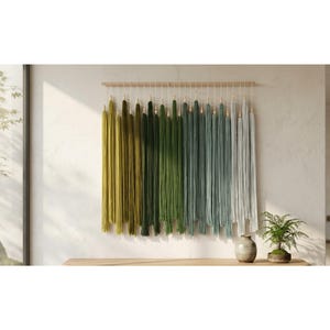 Puede incluir: Un tapiz de pared compuesto por numerosos hilos verticales suspendidos de una varilla de madera horizontal. El hilo presenta un degradado de color que va desde el verde oliva y verde bosque a la izquierda hasta el verde azulado y gris claro a la derecha.