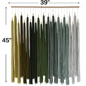 Puede incluir: Un tapiz de pared con una barra de madera horizontal y numerosos hilos verticales en un degradado de color que va del verde oliva al negro, verde oscuro, verde azulado y gris claro. Cada hilo tiene una peque&ntilde;a cuenta de madera. Mide 99 cm de ancho por 114 cm de alto.