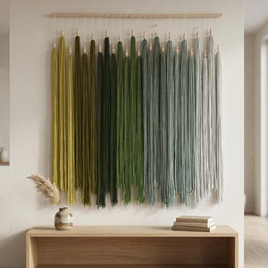 Puede incluir: Un tapiz de pared que presenta una varilla de madera horizontal con m&uacute;ltiples hilos verticales de lana en una paleta de colores degradados. Los tonos transicionan desde el verde oliva y verde bosque al salvia, azul gris&aacute;ceo y gris claro, con cuentas.