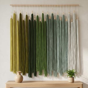 Puede incluir: Un tapiz de pared que presenta una varilla de madera horizontal con m&uacute;ltiples hilos verticales de lana en un degradado de color, que transita de verde oliva y verde bosque a salvia, gris claro y blanco, con peque&ntilde;as cuentas de madera.