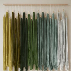 Puede incluir: Un tapiz de pared con una barra de madera horizontal y m&uacute;ltiples hilos verticales de lana en un degradado de colores que van del verde oliva al salvia y gris claro. Cada hilo de lana est&aacute; decorado con una peque&ntilde;a cuenta de madera.
