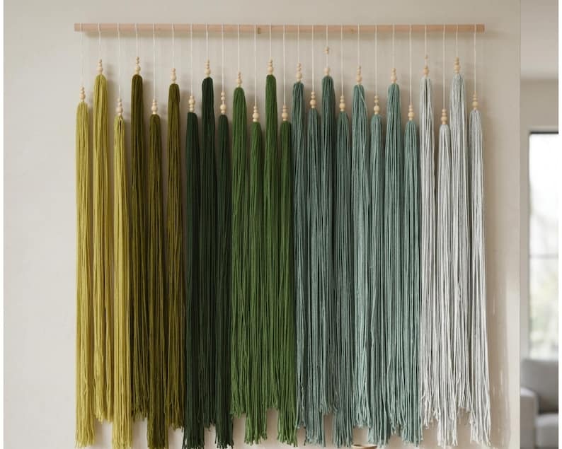 Puede incluir: Un tapiz de pared con una barra de madera horizontal y m&uacute;ltiples hilos verticales de lana en un degradado de colores que van del verde oliva al salvia y gris claro. Cada hilo de lana est&aacute; decorado con una peque&ntilde;a cuenta de madera.