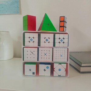 Könnte beinhalten: Eine Sammlung farbenfroher Puzzle-Würfel, darunter ein rot-grüner Würfel, eine grüne Pyramide und mehrere weiße Würfel mit blauen und grünen Akzenten, angeordnet in einem weißen, mehrstufigen Organizer. Die Würfel werden auf einer weißen Oberfläche präsentiert.