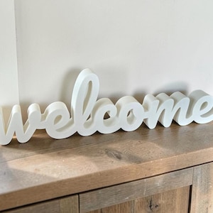Könnte beinhalten: Ein freistehendes weißes Schild mit dem Wort welcome in einer kursiven Kleinbuchstabenschrift. Die Buchstaben sind dick, dreidimensional und haben eine glatte, matte Oberfläche. Das Schild ist so konzipiert, dass es auf einer ebenen Fläche steht.