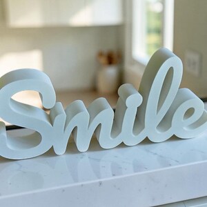 May include: A freestanding white decorative sign featuring the word Smile in a cursive script font. The letters are thick and solid with a smooth matte finish, designed to stand upright on a flat surface. The object is white with soft shadows.