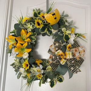 May include: A circular wreath with yellow sunflowers, lilies, and white roses. The wreath has a large bow with a sunflower design and a ceramic bee. Green and yellow foliage surrounds the flowers.