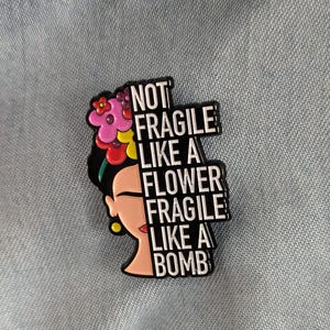 Peut inclure: Une broche en émail métallique représentant le portrait stylisé d'une personne aux cheveux noirs ornés de fleurs roses, rouges et jaunes. Le texte NOT FRAGILE LIKE A FLOWER FRAGILE LIKE A BOMB est imprimé en lettres blanches sur fond noir.