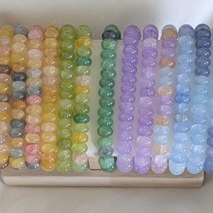 Peut inclure: Une collection de bracelets de perles aux couleurs de l'arc-en-ciel, dont rouge, rose, jaune, vert, violet, bleu et gris. Les bracelets sont présentés sur un présentoir en bois clair, mettant en valeur la variété des perles.