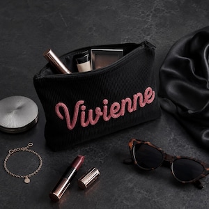 May include: A black corduroy makeup bag with the name "Vivienne" embroidered in pink script. The bag is open, revealing makeup items. A gold bracelet, lipstick, and tortoiseshell sunglasses are also displayed.