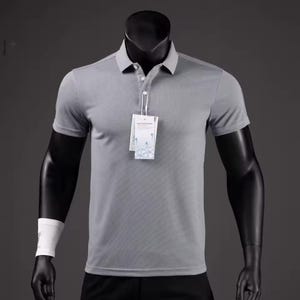 Può includere: Una polo grigia con maniche corte e scollo a camicia. La camicia ha una paramontura a tre bottoni e un'etichetta attaccata. La camicia è esposta su un manichino nero. Un polsino bianco è sul braccio sinistro del manichino.
