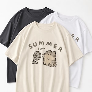 Op de afbeelding: Drie T-shirts met korte mouwen in zwart, crème en wit. Het crèmekleurige shirt heeft een afbeelding van een cartoonkat met een ventilator en de woorden "SUMMER fun". Op het witte shirt staat het woord "ER".