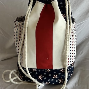May include: A drawstring backpack with a patriotic design. The bag features a central red stripe flanked by white panels, and is accented with navy blue fabric patterned with red and white stars. White rope serves as the drawstring.