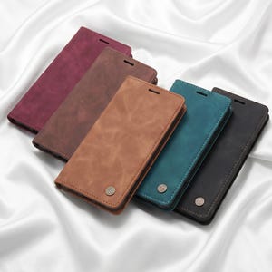 May include: A collection of five rectangular smartphone wallet cases in burgundy, brown, tan, teal, and black. Each case features a smooth, matte finish and a small, circular metallic emblem with a letter T logo near the bottom edge.