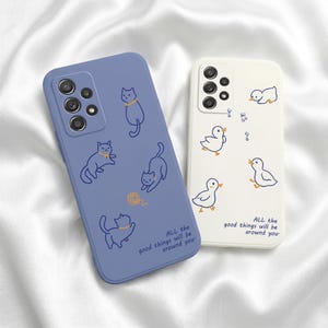 Könnte beinhalten: Zwei Handyhüllen mit Cartoon-Illustrationen. Eine ist periwinkleblau mit Katzenzeichnungen und dem Text "ALL the good things will be around you." Die andere ist cremeweiß mit Entenzeichnungen und demselben Text.