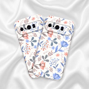 May include: Two white smartphone cases featuring a repeating pattern of small flowers and leaves in shades of light blue, soft peach, and muted gray. Each case has a horizontal camera cutout at the top with circular openings for lenses.