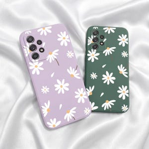 Puede incluir: Dos fundas para teléfono con diseños de margaritas. Una es lavanda, la otra verde oscuro. Ambas tienen margaritas blancas con centros amarillos y tallos marrones. Las fundas son de material liso y tienen recortes para la cámara.