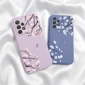 以下が含まれることがあります： 2つの携帯電話ケース。1つはラベンダー色、もう1つはペリウィンクル色で、それぞれに花の模様が施されています。ラベンダー色のケースにはピンクの花と蝶が、ペリウィンクル色のケースには白い花と蝶が描かれています。どちらのケースにもカメラ用の切り抜きがあります。