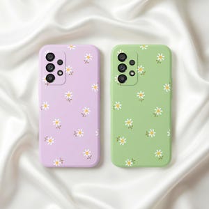 Puede incluir: Dos fundas para teléfono, una lila y otra verde, cada una adornada con un patrón repetido de pequeñas margaritas blancas con centros amarillos y tallos verdes. Las fundas tienen un acabado liso y un módulo de cámara.
