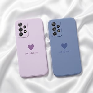 May include: Two phone cases, one lavender and one blue, are displayed on a white satin surface. Each case features a heart graphic and the text "be loved~". The cases have a matte finish and are designed for a smartphone.