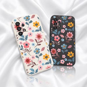 May include: Two smartphone cases featuring a floral pattern. One case is cream-colored with pink, yellow, and blue flowers and green leaves. The other case is black with the same floral design. Both cases have a cutout for a multi-lens camera system.