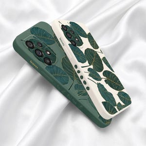 May include: Two mobile phone cases, one in dark green and one in off-white, both featuring a repeating pattern of dark green leaves with gold-colored veins. Each case has a camera cutout and the word leaves printed on the side edge.