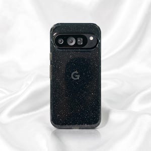 Op de afbeelding: Een zwarte smartphonehoes met een fijne glitterafwerking en een transparante rand. De hoes heeft een horizontale uitsparing voor de camerabalk aan de bovenkant met drie ronde lensopeningen en een flitser. Een kleine letter G staat in het midden.