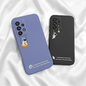 May include: A periwinkle blue and a matte black phone case, each featuring a camera cutout array. The blue case shows an astronaut on a crescent moon, while the black case shows an astronaut reaching for a star. Both include the text: THE ASTRONAUTS PILOTED THEIR CRAFT DOWN TO THE LUNAR SURFACE.