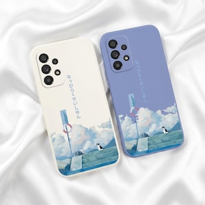 Pode incluir: Duas capas para smartphone, uma creme e uma azul-pervinca, cada uma com uma ilustração impressa de um gato pequeno em degraus de pedra perto de uma boia, nuvens e texto japonês. As capas têm aberturas para o sistema de câmara.