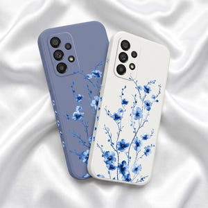 May include: Two smartphone cases, one in matte periwinkle blue and one in white, both featuring a delicate watercolor-style illustration of thin, dark blue branches with small, vibrant blue flowers extending across the back surface.