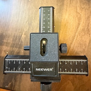 May include: A black Neewer macro focusing rail with a central mounting plate featuring a textured surface and a metal screw. The device includes two perpendicular sliding tracks with white printed scales marked from 1 to 6 inches for precise adjustment.