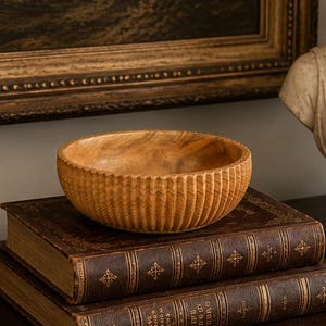 May include: A light brown wooden bowl with a ribbed exterior texture sits atop two stacked leather-bound books. The scene includes two brass candlesticks, a magnifying glass with a metal handle, and a white stone bust of a person on a dark base.