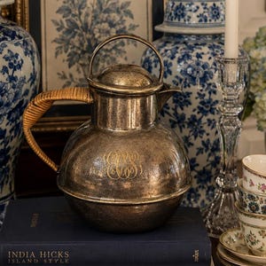 May include: A silver-toned metal pitcher with a woven rattan handle and a monogrammed front, resting on two stacked books titled India Hicks Island Style and A Privileged Life, surrounded by blue and white porcelain jars and floral teacups.