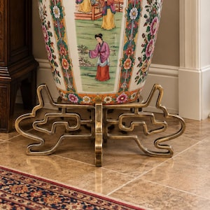 May include: A large, ornate porcelain floor vase featuring colorful hand-painted floral patterns and detailed scenes of figures in traditional attire. The vase has a wide, flared rim and rests on a decorative, gold-toned metal stand.