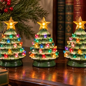 May include: Three ceramic tabletop Christmas trees in a glossy green finish with white snow-like accents on the branches. Each tree is decorated with small, multi-colored glowing bulbs and topped with a bright, illuminated yellow star.