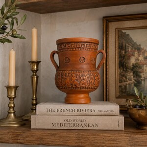 May include: A terracotta-colored vase with two handles, featuring a detailed relief of figures and patterns. The vase sits atop two books with the titles "The French Riviera" and "Old World Mediterranean". Flanking the vase are brass candle holders and a framed painting.