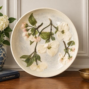 May include: A round ceramic bowl with a cream-colored base featuring a raised, three-dimensional relief of white dogwood flowers, green leaves, and brown branches. The interior surface has a subtle, mottled light blue and white glaze finish.