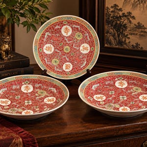 May include: Three round porcelain plates featuring a vibrant red background with intricate gold and white floral patterns, green geometric borders, and four white circular medallions containing black Chinese characters arranged symmetrically.