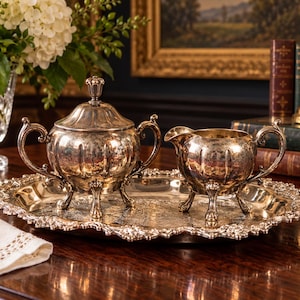 May include: A silver-plated sugar bowl with a lid and a matching creamer pitcher, both featuring ornate handles and feet, arranged on an oval silver-plated serving tray with a decorative scalloped edge and intricate embossed detailing.